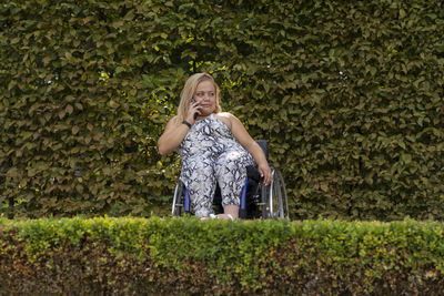 Young woman on wheelchair talks on phone in green park at summer day. busy female adult with