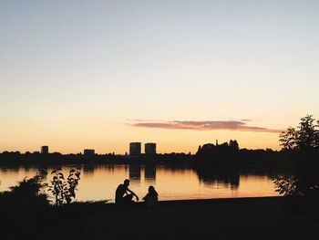 Silhouette people sitting on riverbank against clear sky