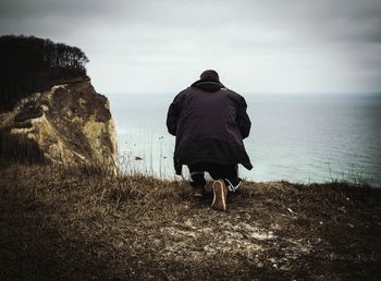 Rear view of man crouching on mountain by sea against sky