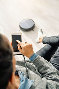 From above of crop person in casual clothes using mobile app and controlling robotic vacuum cleaner while sitting in room with light wooden floor