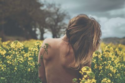 Rear view of shirtless young woman standing by flowers against cloudy sky
