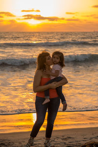 Grandmother kissing granddaughter at beach against cloudy sky during sunset