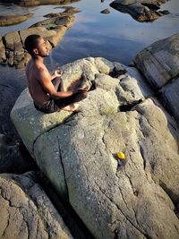 Side view of shirtless young man meditating while sitting on rock at beach