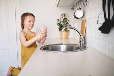 European child of 4 years old washes glass in sink on his own, bright kitchen in real interior, 