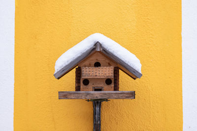 Close-up of wooden birdhouse on yellow wall. snow covered roof.