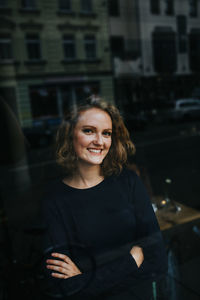 Portrait of smiling young woman with arms crossed seen through glass window