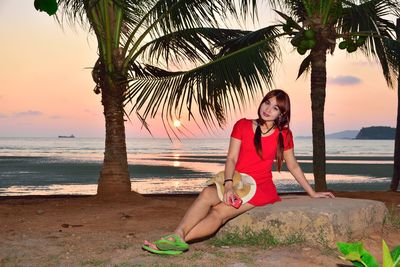 Full length portrait of young woman in red dress sitting on rock at beach