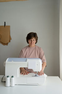 Concentrated female dressmaker in eyeglasses working on sewing machine at table with threads in light professional workshop with special equipment