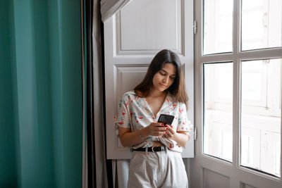 Calm brunette female browsing on smartphone standing near window at home