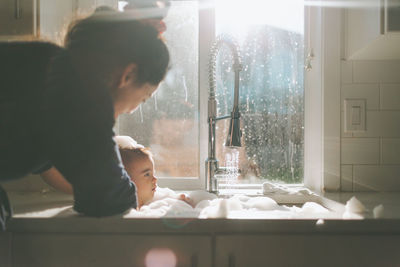 Mother bathing son in kitchen sink by window at home
