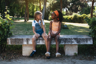 African american girl smiling and best friend sitting together on bench in green park in sunny day