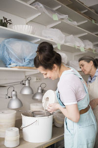 Senior female potter looking at young employee pouring clay from vase in bucket at workshop