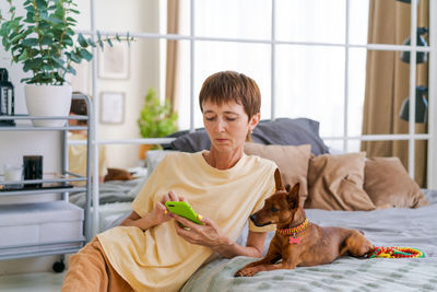 Mature woman with a phone in her hand with a pygmy pinscher in the bedroom