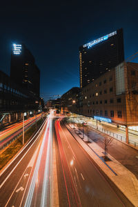 Light trails on city street by buildings against sky at night