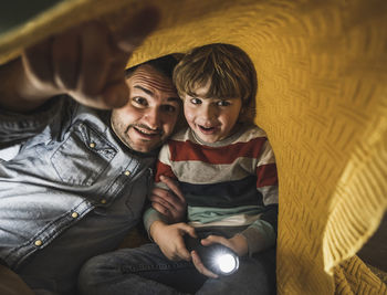 Happy father gesturing with son holding torch under yellow blanket