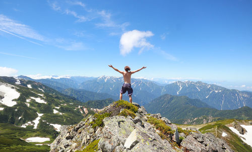 Rear view of shirtless man with arms outstretched standing on mountain against sky