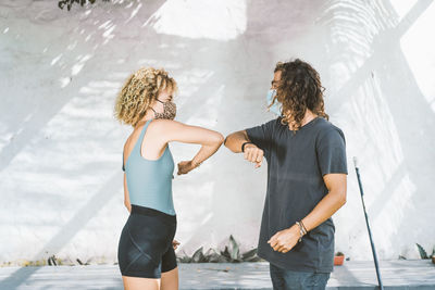 Man and woman wearing mask giving elbow bump while standing against wall