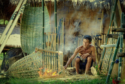 Shirtless senior man sitting by fire outside hut