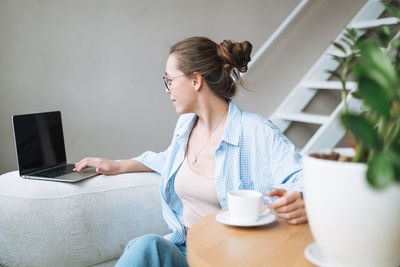 Young woman in blue shirt using laptop drinking coffee in room at home