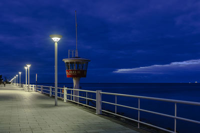 Illuminated street lights by sea against sky at dusk