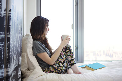 Side view of woman with coffee cup looking away while sitting on bed at home