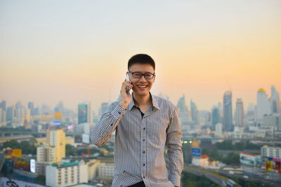 Businessman talking on smart phone while standing against cityscape during sunset