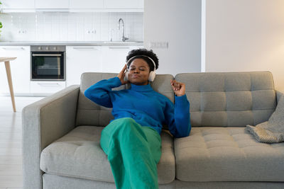Carefree cheerful african american woman listening to music holding headphones to ears sits on sofa