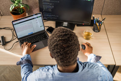 Black man writing lines of code on desktop pc with two monitors and a laptop aside in stylish office