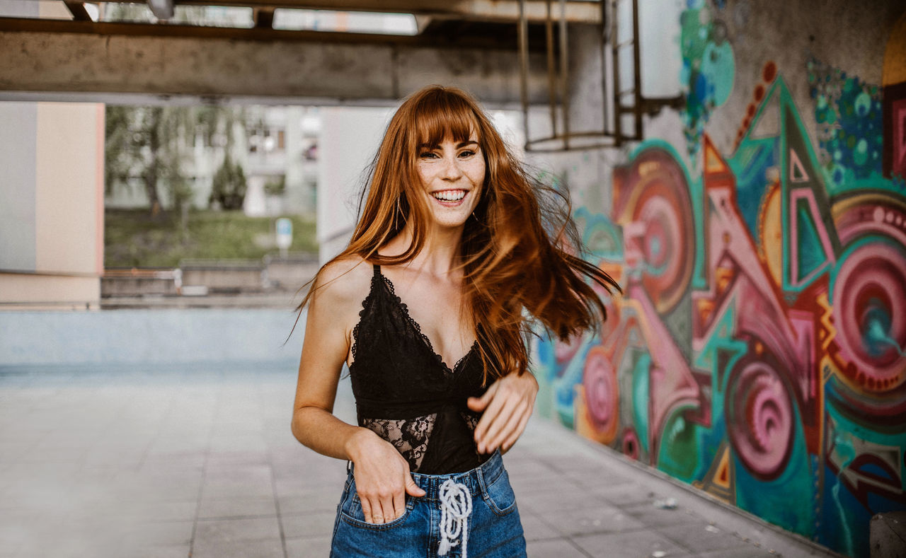 young adult, young women, long hair, one person, hairstyle, casual clothing, hair, front view, smiling, three quarter length, happiness, graffiti, standing, architecture, real people, emotion, portrait, leisure activity, lifestyles, beautiful woman, fashion, outdoors