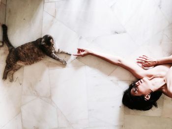 High angle view of naked woman lying on floor while pointing towards tabby cat at home