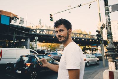 Portrait of young man holding mobile phone while standing on sidewalk in city