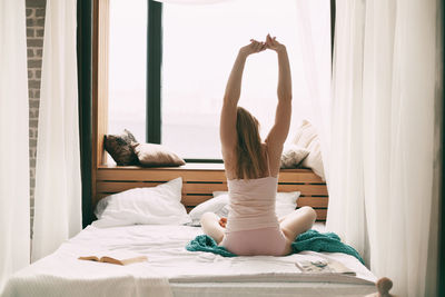 A beautiful girl sits on the bed in the early morning and stretches after waking up, 