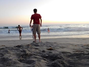 Rear view of man standing at beach against sky during sunset