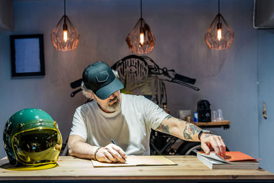 Front view of a man writing in a notebook on a wooden table next to a motorcycle helmet person