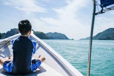 Rear view of boy sitting on boat deck in sea against sky