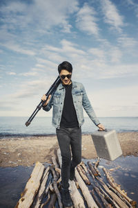 Full length of young man with briefcase and tripod walking on logs at beach