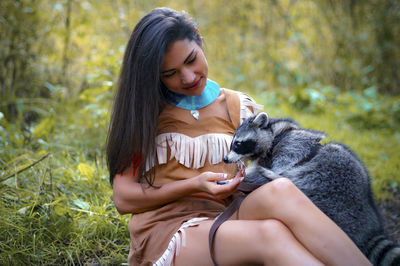 Smiling young woman in traditional clothing sitting by raccoon in forest