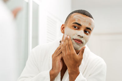Young man with facial mask looking in mirror at home