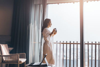 Side view of young woman looking through window at home