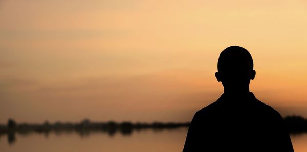 Silhouette man standing by lake against sky during sunset