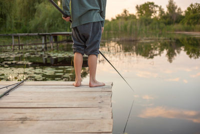 Low section of boy fishing in lake while standing on pier during sunset