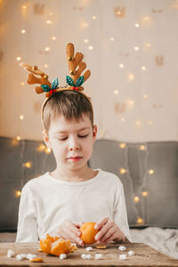 Boy in christmas antlers, sits and peels a tangerine on the background of a yellow garland.