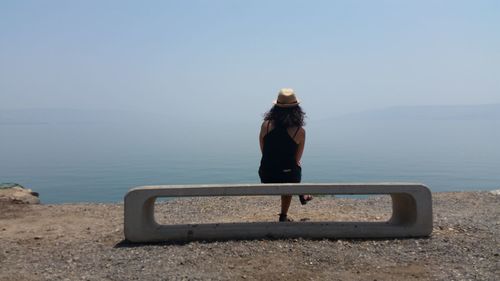 Rear view of woman sitting on concrete seat by lakeshore against sky