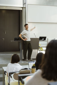 Male professor standing with hand on hip while teaching students in lecture hall at university