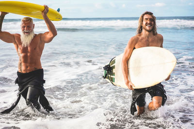 Shirtless men holding surfboards while walking in sea