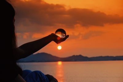 Woman holding crystal ball against sea during sunset