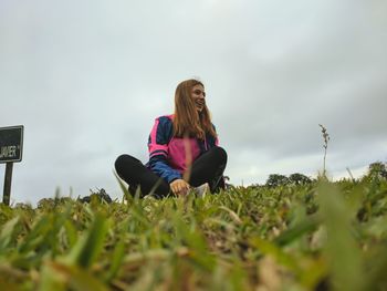 Low angle view of young woman sitting on grass against sky