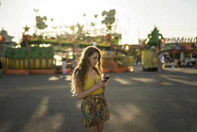 Female with wavy hair standing at fairground and browsing mobile phone at sundown in summer