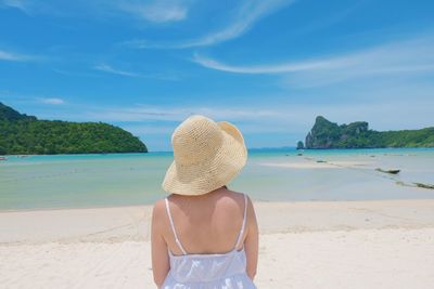 Rear view of woman wearing hat while standing at beach against blue sky during sunny day