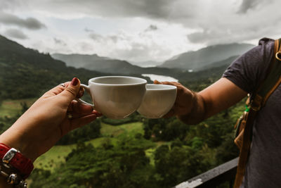 Cropped hands of couple toasting coffee cups against mountain